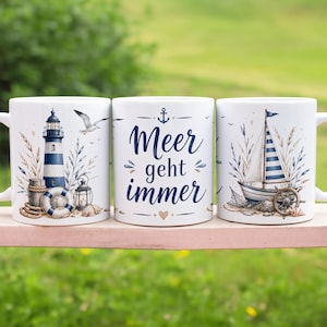 Könnte beinhalten: Ein weißer Keramikbecher mit nautischem Motiv, bestehend aus einem blau-weißen Leuchtturm, einem Segelboot, einem Rettungsring und dem blauen Schriftzug "Meer geht immer", verziert mit kleinen Ankern, Herzen und Küstenpflanzen.