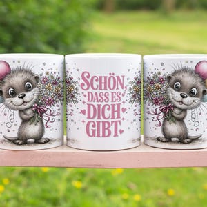 May include: Three white ceramic mugs with a cartoon otter holding flowers and balloons. The otter is gray and white with a smiling face. The middle mug has the text "Sch&ouml;n dass es dich gibt" in pink.