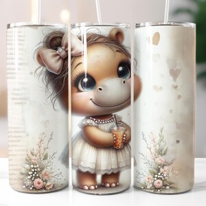 Könnte beinhalten: Ein weißer Becher mit einem braunen und weißen Cartoon-Nilpferd, das ein weißes Kleid trägt und ein rosa Getränk hält. Das Nilpferd hat eine rosa Schleife im Haar und blaue Augen. Der Becher hat ein florales Design am Boden.