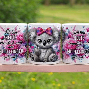 Puede incluir: Tres tazas de cerámica blanca con un diseño floral y un koala de dibujos animados. Las tazas tienen el texto "Ich bin HEUTE ziemlich nah am MITTELFINGER gebaut!" impreso en ellas.