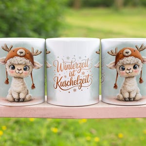 May include: Three white ceramic mugs with a cute cartoon llama wearing a brown hat with antlers. The mugs have the text "Winterzeit ist Kuschelzeit" in brown script. The mugs are on a pink surface with a green background.