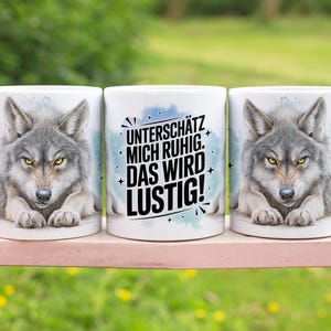 May include: Three white ceramic mugs with a wolf design. The wolf has gray fur and yellow eyes. The middle mug has the text "UNTERSCHÄTZ MICH RUHIG. DAS WIRD LUSTIG!" in black and white.