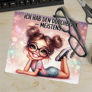 Puede incluir: Un paño de limpieza de microfibra con una ilustración de dibujos animados de una niña con gafas, con el texto alemán "ICH HAB DEN DURCHBLICK - MEISTENS." Gafas negras y tijeras sobre la mesa.