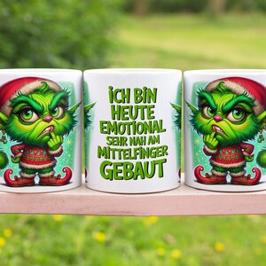 May include: Three white ceramic mugs with a cartoon Grinch design. The Grinch is green with a red Santa hat and a red and white sweater. The middle mug has the German text "Ich bin heute emotional sehr nah am Mittelfinger gebaut."