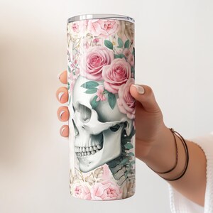 Pu&ograve; includere: Un bicchiere in acciaio inossidabile bianco con un design floreale che presenta un teschio con rose rosa e foglie verdi.