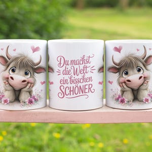 Op de afbeelding: Drie witte keramische mokken met een schattig cartoon koeienontwerp. De koe heeft een lichtbruine vacht, roze accenten en is omringd door roze hartjes en bloemen. De middelste mok heeft de tekst "Du machst die Welt ein bisschen SCHÖNER".