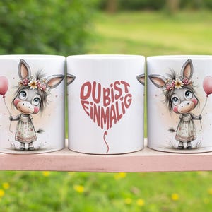May include: Three white ceramic mugs featuring a cartoon donkey with a floral crown and a pink heart balloon. The central mug has the German text "DU BIST EINMALIG" in a heart-shaped balloon design.