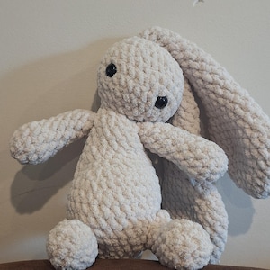 Może przedstawiać: Pluszowa zabawka kr&oacute;liczka wykonana na szydełku z długimi, zwisającymi uszami. Kr&oacute;liczek siedzi na brązowej powierzchni i jest wykonany z miękkiej, białej przędzy.