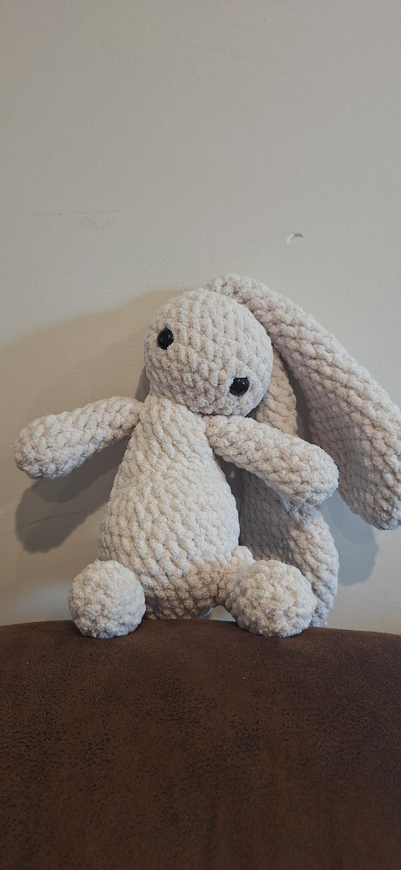 Może przedstawiać: Pluszowa zabawka kr&oacute;liczka wykonana na szydełku z długimi, zwisającymi uszami. Kr&oacute;liczek siedzi na brązowej powierzchni i jest wykonany z miękkiej, białej przędzy.