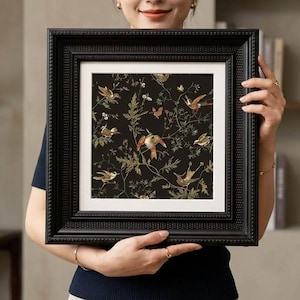 May include: A framed artwork with a detailed illustration of birds, butterflies, and foliage in brown, green, and gold against a black background. The artwork is set within a white mount and a black, ornate frame.