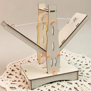 May include: A light-colored wooden mechanical model featuring a central vertical support structure with cutouts, two angled arms extending outward, internal white gears, orange fasteners, and a thin white string connecting the two arms.