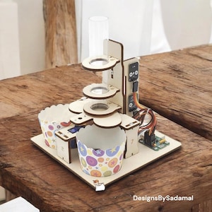 Peut inclure: Un distributeur de cupcakes en bois avec un tube en plastique transparent et un petit circuit imprimé. Le distributeur a trois supports à cupcakes avec des doublures en papier colorées. Le distributeur est sur une table en bois.