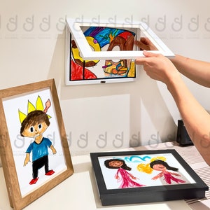 May include: Three framed children's drawings, one with a boy wearing a crown, another with two girls, and a third with abstract shapes and colors. The drawings are hanging on a white wall and one is being held by a person's hand.