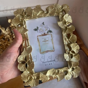 May include: Gold-colored photo frame with a decorative design of leaves and flowers. The frame is 4 inches by 6 inches and has a white mat.