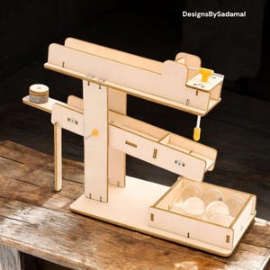 Könnte beinhalten: Ein Holzspielzeug mit einer Rampe und einem Hebel, der einen kleinen Ball von einer Ebene zur nächsten bewegt. Das Spielzeug ist aus Naturholz gefertigt und hat ein schlichtes Design. Der Ball ist aus klarem Glas und im unteren Fach sichtbar.