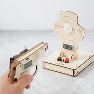 Può includere: Una pistola giocattolo in legno chiaro con display digitale e caratteri cinesi, abbinata a un supporto per bersaglio in legno con un centro circolare, uno schermo per il punteggio digitale, un cicalino nero e un pulsante rosso.
