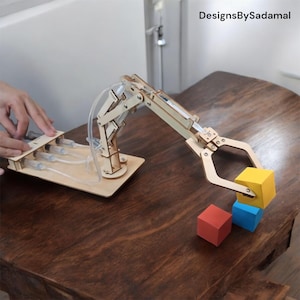 May include: A wooden robotic arm with a claw-like hand is shown picking up a yellow block. The arm is connected to a base with three syringes. The arm is on a wooden table with two other blocks, one red and one blue.