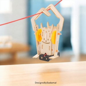 May include: A wooden toy robot with yellow gears and a red string attached to its arms. The robot is suspended in the air above a wooden surface.