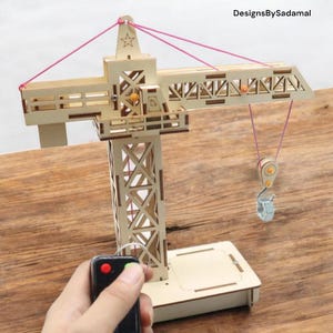 May include: A wooden toy crane with a red and white star on top, a red remote control, and a hook hanging from the crane's arm. The crane is on a wooden base.