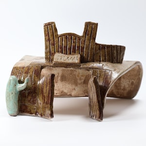 May include: A brown ceramic sculpture depicting a saloon building with a curved roof and a small light blue cactus figure attached to the front left side. The structure features a sign with the word SALOON embossed on the front facade.