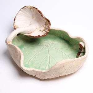 May include: A ceramic dish shaped like a leaf with a light green interior featuring embossed veins. A white mushroom with a brown-edged cap is attached to the rim, and a small brown creature figure is perched on the opposite side of the dish.