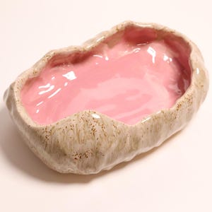 May include: An oval ceramic bowl with a glossy pink interior and a textured, beige and brown marbled exterior. The rim has an irregular, organic shape with a wavy edge, and the overall surface has a smooth, reflective finish throughout.