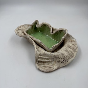 May include: A handcrafted ceramic dish set. The set features a large, organically shaped base with a smaller, leaf-shaped dish nested within. The interior of the leaf-shaped dish is painted a bright green, contrasting with the speckled, off-white exterior.