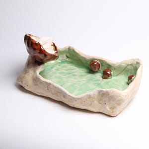 May include: A handmade ceramic tray with an irregular, organic shape and a textured off-white exterior. The interior features a glossy light green glaze with a leaf-like pattern, accented by a brown sculpted mushroom and three small brown spheres.