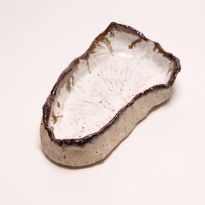 May include: An irregular, hand-formed ceramic dish with a textured, speckled beige exterior and a smooth, glossy white interior. The rim is defined by a dark, metallic brown glaze that drips slightly down the sides, creating a rustic appearance.