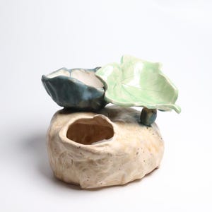 May include: A ceramic sculpture featuring a textured, beige, rock-like base with a hollow opening. Attached to the top are two distinct leaf-shaped bowls, one finished in a glossy dark blue glaze and the other in a light, pale green glaze.