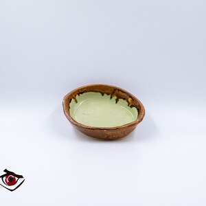 Può includere: Una piccola ciotola in ceramica di forma ovale con interno smaltato verde chiaro e bordo esterno testurizzato marrone scuro. La ciotola presenta un bordo organico irregolare e una finitura interna liscia e lucida, in contrasto con l'aspetto terroso e rustico del bordo esterno.