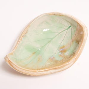 May include: A small, leaf-shaped ceramic dish with a glossy finish. The interior features a pale green glaze with a visible leaf vein pattern and subtle brown accents. The outer rim is a smooth, light beige colour, creating a natural aesthetic.