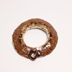 May include: A circular ceramic ring with a textured brown surface featuring a grid-like pattern. The inner edge is glazed in white, and the outer surface has a glossy finish. Three small, dark brown, rounded protrusions are attached to the bottom.