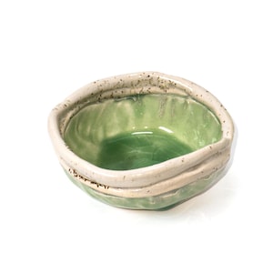 Puede incluir: Cuenco de cerámica pequeño, hecho a mano, con interior verde claro y borde texturizado blanquecino. El cuenco tiene una forma orgánica única y está sobre un fondo blanco. El borde tiene pequeñas motas oscuras.