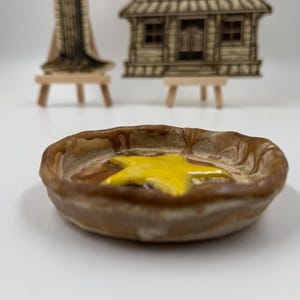 Könnte beinhalten: Eine kleine, runde Keramikschale mit brauner Außenseite und einem leuchtend gelben Stern in der Mitte. Die Schale hat einen leicht gewellten Rand. Der Hintergrund umfasst Miniatur-Holzstrukturen auf kleinen Staffeleien.