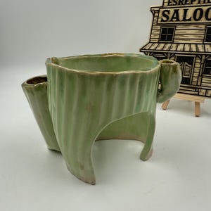 May include: A light green ceramic shaving scuttle with a ribbed exterior. The scuttle has a handle and a side compartment for a shaving brush. The base is open, and the piece is on a white surface.