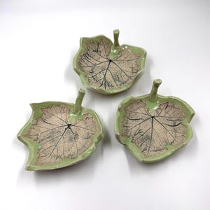 May include: Three ceramic leaf-shaped dishes with a green glaze and a raised vein pattern. The dishes have a small handle on the top.
