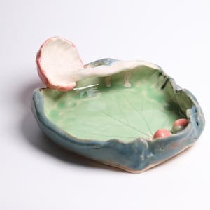 May include: A handmade ceramic trinket dish with an irregular, organic shape. The interior is glazed in a light green with a leaf vein pattern, while the exterior is a muted blue. A sculpted pink mushroom extends from the rim into the centre.