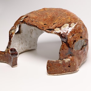 May include: A ceramic cave-like structure with a glossy, mottled brown and rust-coloured exterior and a textured, speckled white interior. The object features an arched opening and irregular, organic edges with small, raised dark brown nodules.