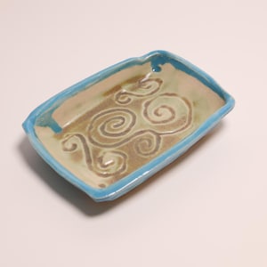 Pode incluir: Uma bandeja retangular de cerâmica com uma borda azul-clara brilhante e uma base interna bege. O centro da bandeja apresenta uma série de padrões de espirais em relevo num tom castanho suave, criando uma superfície texturizada no prato.