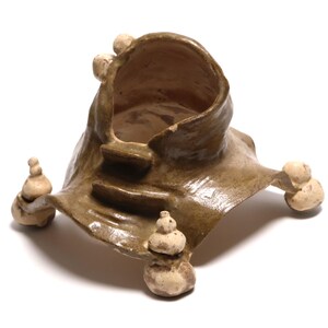 Może przedstawiać: Ręcznie wykonana rzeźba ceramiczna o unikalnym, organicznym kształcie. Element posiada centralną strukturę przypominającą misę ze schodami, wspartą na rozszerzonej podstawie i zaokrąglonych, ułożonych w stos akcentach. Kolor to ciepły, ziemisty brąz.