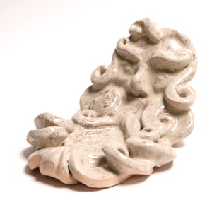 Könnte beinhalten: Eine cremefarbene Keramik-Wandskulptur, die ein stilisiertes Gesicht mit fließenden, gelockten Haar- und Bartdetails zeigt. Die Oberfläche hat ein glänzendes Finish mit dezenten braunen Sprenkeln, die der gesamten Form ein strukturiertes Aussehen verleihen.