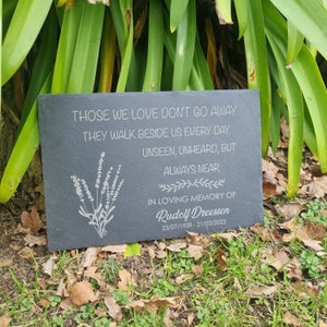 May include: A black slate memorial plaque with a white inscription that reads "Those we love don't go away. They walk beside us every day. Unseen, unheard, but always near. In loving memory of Rudolf Dreessen 23/07/1939 - 21/03/2023". The plaque is decorated with a white silhouette of a bouquet of lavender.