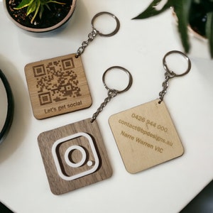 May include: Two wooden keyrings with engraved designs. One keyring features a QR code with the text "Let's get social". The other keyring has a white Instagram logo on a brown background and contact information for a business.