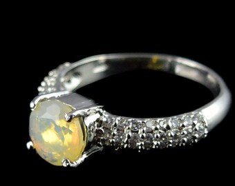 Puede incluir: Un anillo de plata con una gran piedra preciosa de ópalo amarillo y redondo. La banda está adornada con pequeñas piedras preciosas transparentes. El anillo se presenta sobre un fondo negro, resaltando su brillo y diseño.