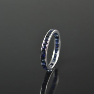 May include: A silver band ring with a row of square-shaped blue sapphires set around the entire band.