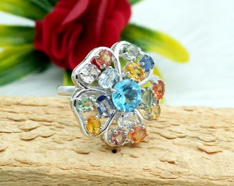 May include: A silver ring with a flower design featuring a large blue gemstone surrounded by smaller multi-colored gemstones.