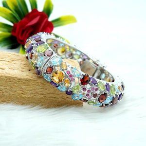 Puede incluir: Un brazalete de plata con una banda ancha incrustada con una variedad de piedras preciosas de colores, incluyendo amatista, citrino, peridoto, granate y aguamarina.