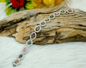 May include: A silver bracelet with oval-shaped gemstones in various colors, including green, pink, and blue. The gemstones are surrounded by smaller, clear stones.