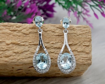 May include: A pair of silver drop earrings featuring oval, light blue gemstones. The earrings have a teardrop design with a halo of small, clear stones around the larger oval stone. The earrings are displayed on a wooden surface.